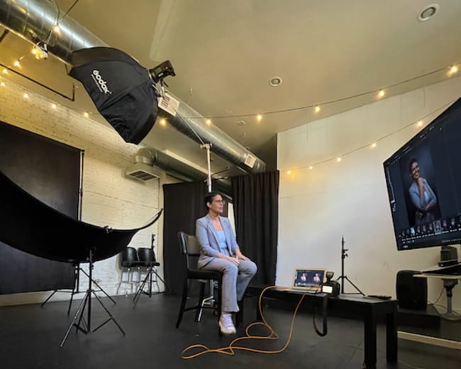 A person in formal attire sits on a chair in a photography studio with soft lighting and setup equipment around them.