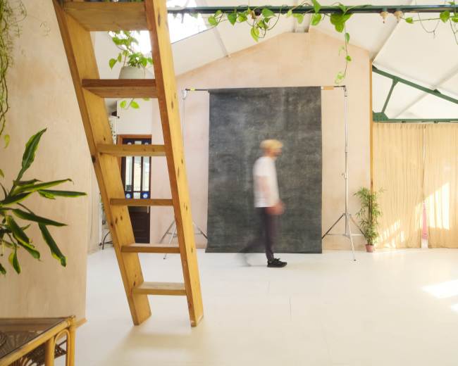 A blurred figure walks past a backdrop in a bright, minimalist studio with wooden stairs and plants.