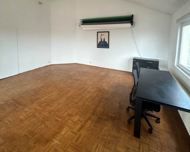 The image shows a spacious, empty room with a wooden floor, a desk and chair, and a wall-mounted backdrop.