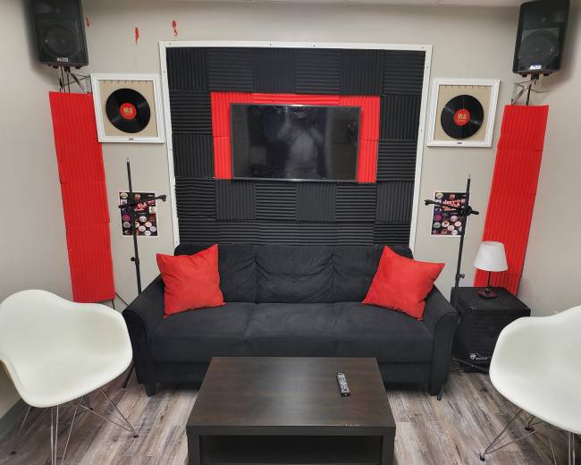 The image shows a modern lounge area with a black couch, red accent pillows, and a wall decorated with black acoustic panels and vintage record displays, alongside a television.