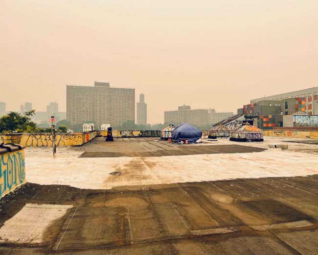 A rooftop scene features scattered tents and graffiti against a backdrop of tall buildings shrouded in a hazy atmosphere.
