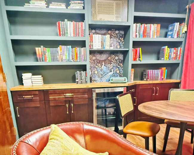 The image shows a cozy reading nook with a leather armchair, a round table, and bookshelves filled with various books in a well-lit space.