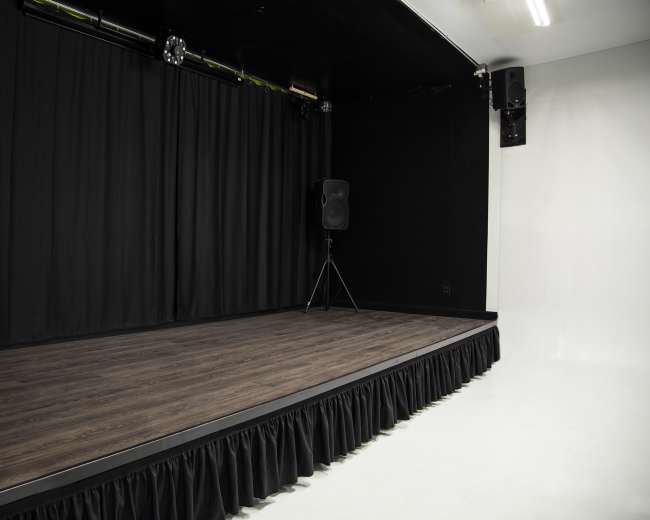 The image shows a stage with a wooden floor and black curtains, featuring speakers on either side.