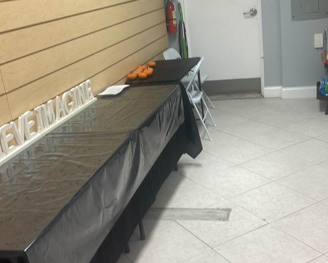 The image shows a room with a light gray wall, a black tablecloth covering a table on the left, and a closed door in the background.