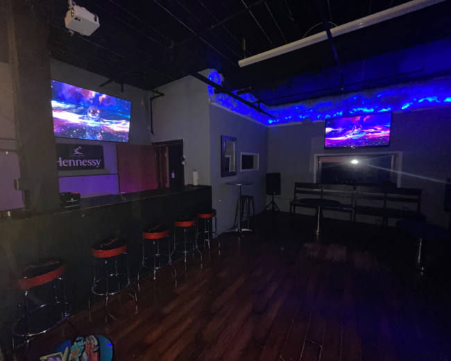 A dimly lit bar area with two wall-mounted screens and a row of red bar stools lined up at the counter.