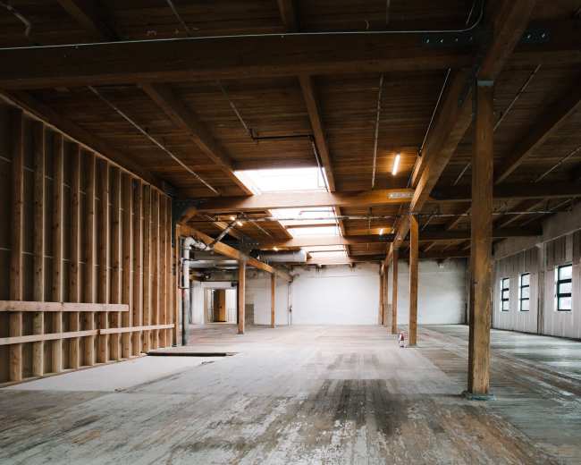 The image shows an empty industrial space with wooden beams, a wooden floor, and an unfinished wall on the left side.