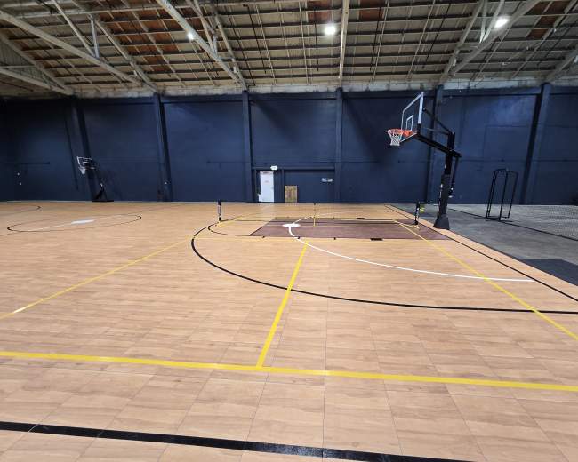 The image shows a spacious indoor basketball court with a wooden floor, marked lines, and a single basketball hoop.