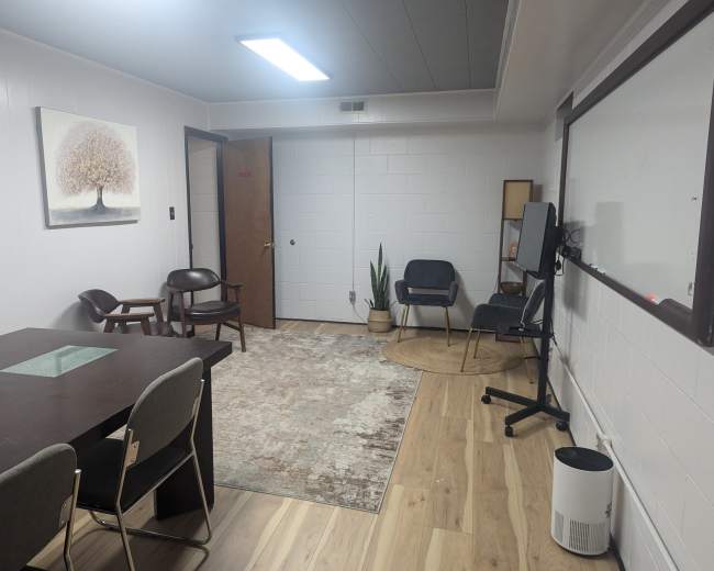 The image shows a minimalist office space with a large area rug, a wooden table and chairs, a whiteboard, and a few decorative plants.