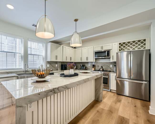 The image shows a modern kitchen featuring white cabinetry, stainless steel appliances, and a large marble-topped island with pendant lighting.