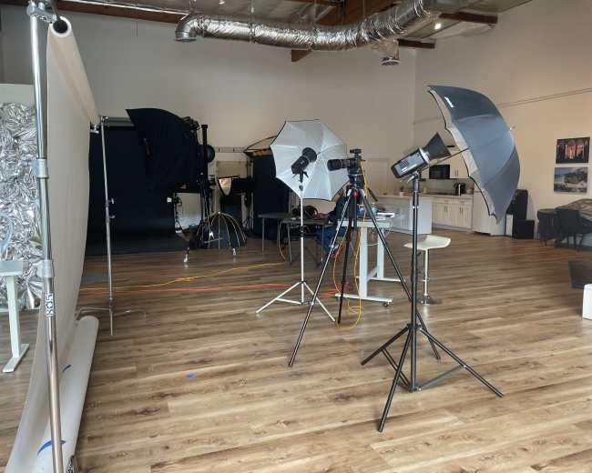 The image shows a photography studio set up with various lighting equipment, backdrops, and a spacious wooden floor.