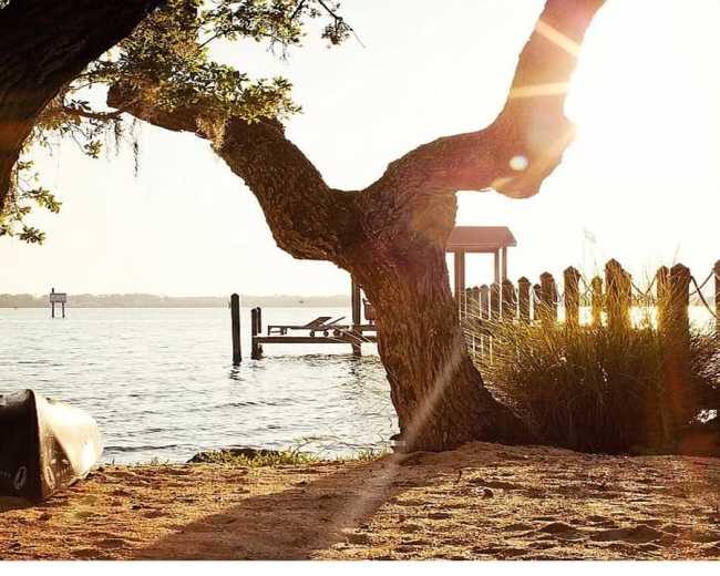 The scene features a sunlit waterfront with a wooden dock, a canoe on the sand, and trees framing the view.