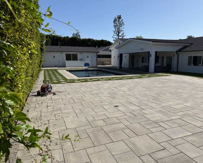The image shows a spacious outdoor area with a swimming pool, surrounded by a neatly designed patio and a grassy border, set against a backdrop of a modern house.