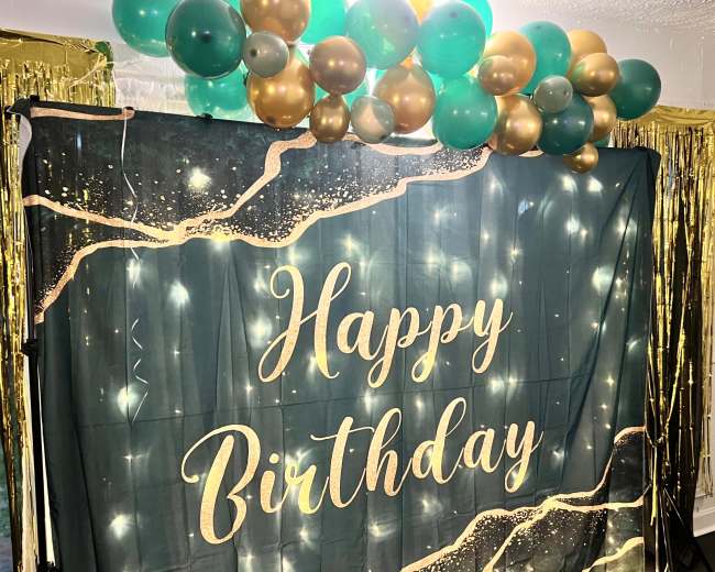 A "Happy Birthday" banner is displayed against a dark fabric backdrop, adorned with gold and teal balloons and string lights.