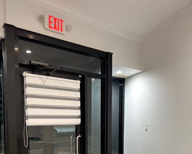 The image shows a modern entryway with a glass door, a roller shade, and a small reception area featuring a white counter beside the exit sign.