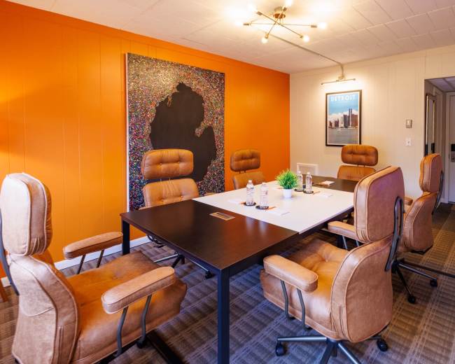 The image shows a modern conference room with a large black table surrounded by brown swivel chairs, an orange accent wall, and artwork featuring a colorful design.