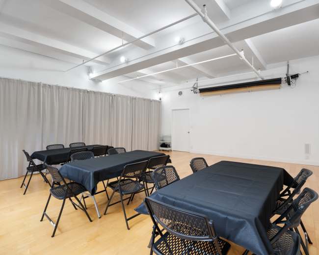 A spacious room is set up with black tables and chairs, divided by gray curtains, and features a plain wall at one end.