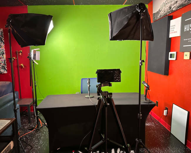 The image shows a video recording setup with a green screen background, a microphone on a table, and studio lights positioned on either side.