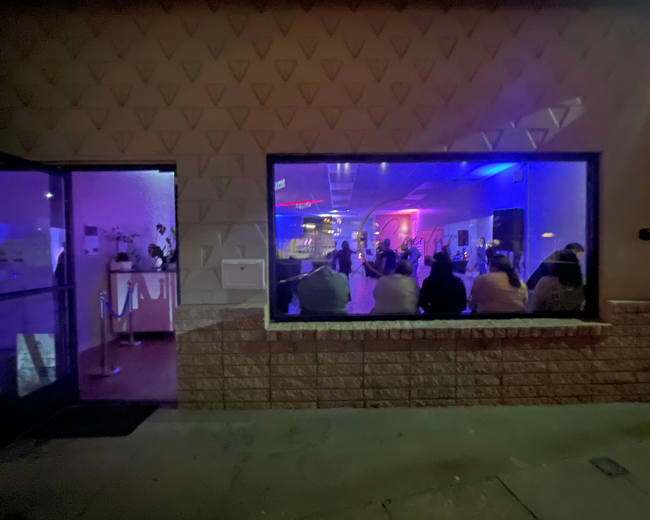 A group of people is seated in front of a brightly lit stage inside a venue, visible through a large window.
