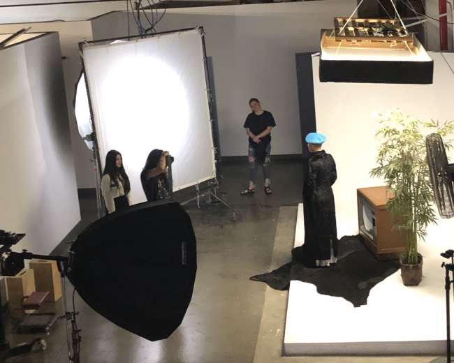 A photoshoot is taking place in a studio, featuring a model in a black outfit with a blue hat, while two photographers and a stylist are positioned nearby.