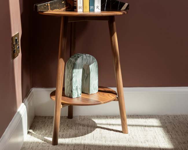 A round wooden side table in a corner holds a stack of books and a decorative object against a brown wall.