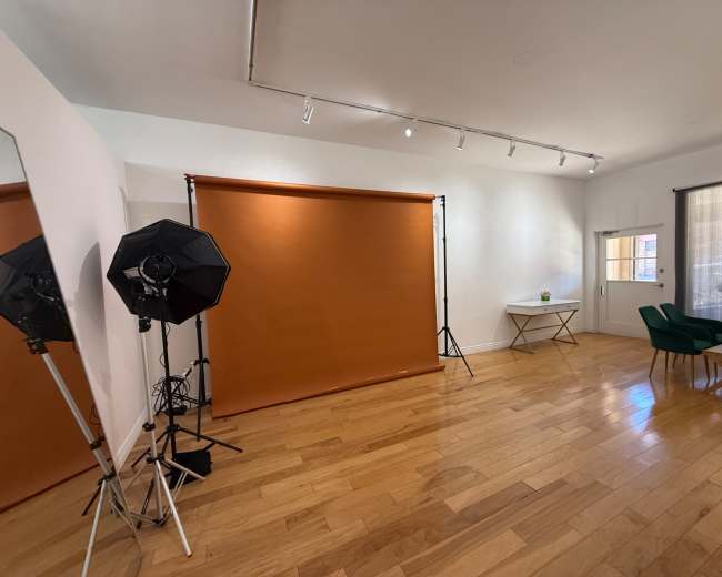 The image shows a well-lit room featuring a brown backdrop set up on stands, two softbox lights, and minimal furnishings including a green chair and a white table.