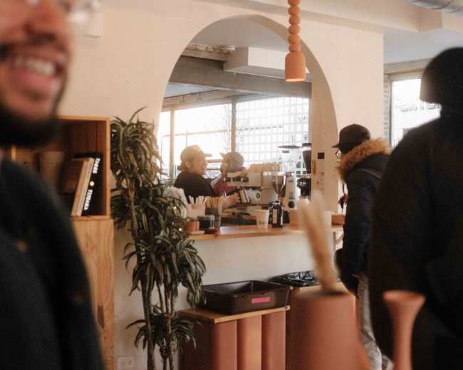 The image shows a cozy café interior with a person smiling in the foreground, while baristas prepare drinks in the background amidst shelves and plants.