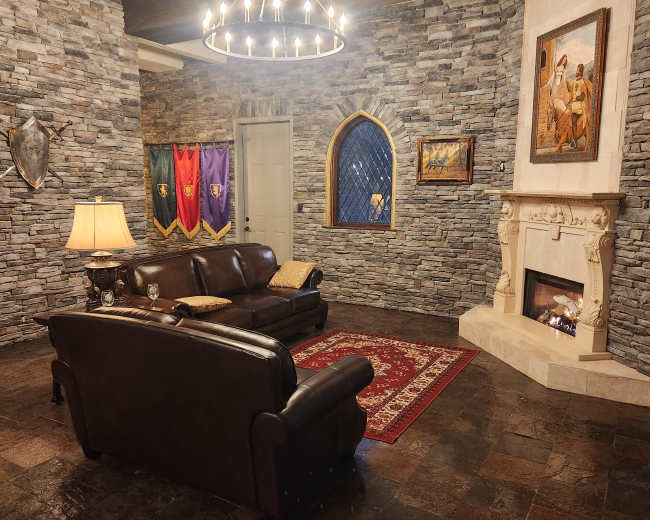 The image shows a cozy living room with stone walls, a fireplace, a dark leather sofa, and medieval-themed decorations.