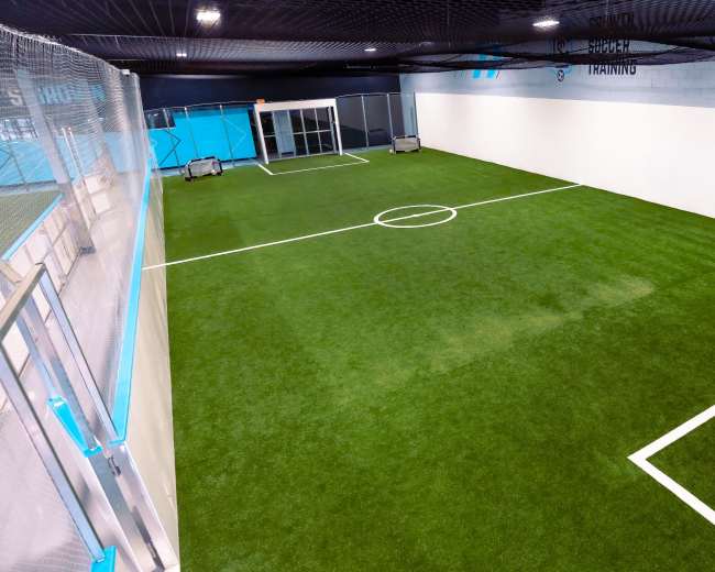 An indoor soccer training facility with artificial turf, marked with boundary lines and goals at each end.