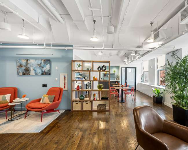 The image shows a modern office space with a mix of orange and brown seating, wooden floors, and shelving displaying decorative items, along with a dining area in the background.