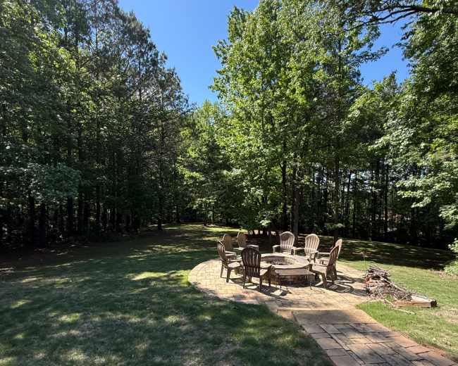 A stone patio with a circular arrangement of wooden chairs sits in a clearing surrounded by trees.
