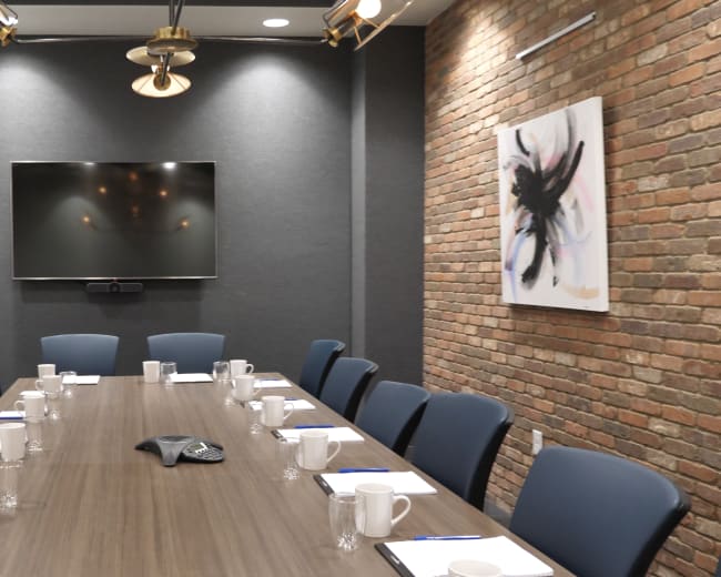 A modern conference room features a long wooden table with chairs, a large wall-mounted screen, a whiteboard, and brick accents.