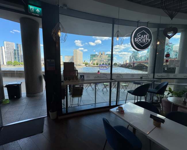 The image shows an indoor café with large windows overlooking a river and city skyline on a sunny day.