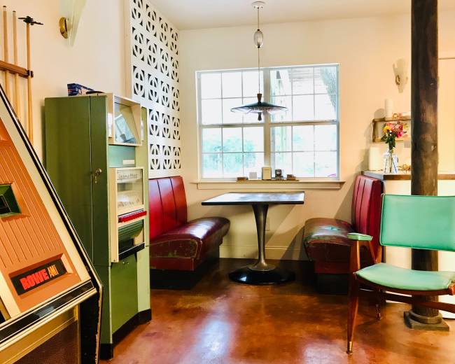 The image shows a dining area with red leather booths, a round table, a retro green refrigerator, and a teal chair, all set against a backdrop of patterned walls and a large window.