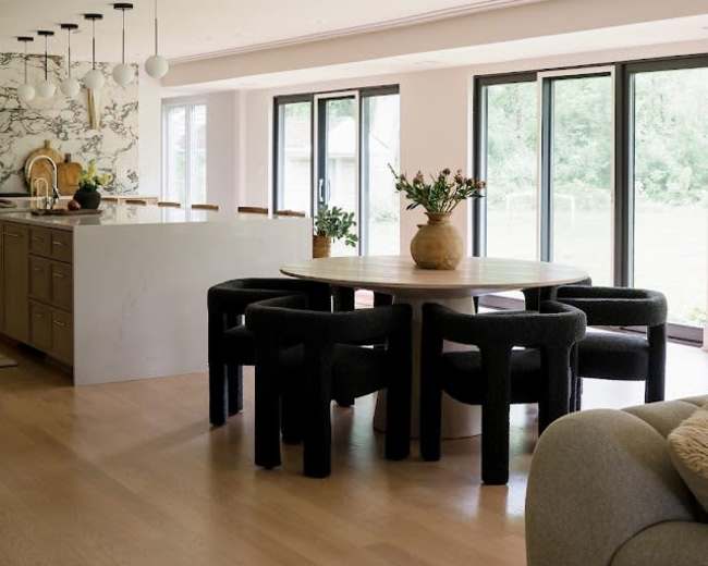 A modern dining area features a circular table surrounded by black upholstered chairs, with large windows providing natural light.