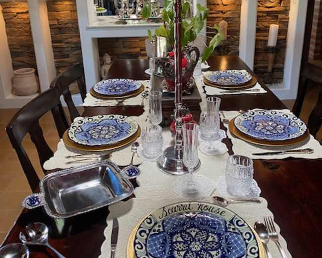 A dining table is set with blue and white patterned plates, glassware, and a centerpiece candle holder, surrounded by stone wall accents in the background.