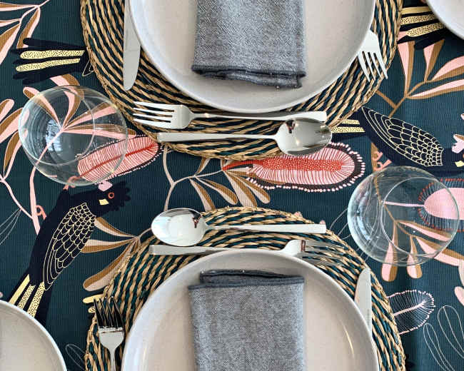 The image shows a dining table set with two plates, gray napkins, cutlery, and glasses, arranged on a patterned table runner.