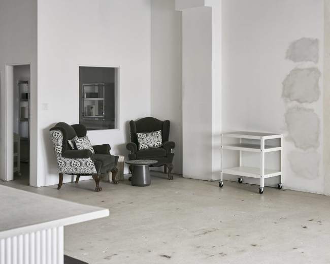 The image shows a minimalist interior space with two upholstered armchairs and a wheeled cart against a blank wall.