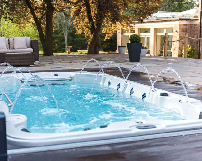 A hot tub with water jets is set on a patio surrounded by trees and a modern home.