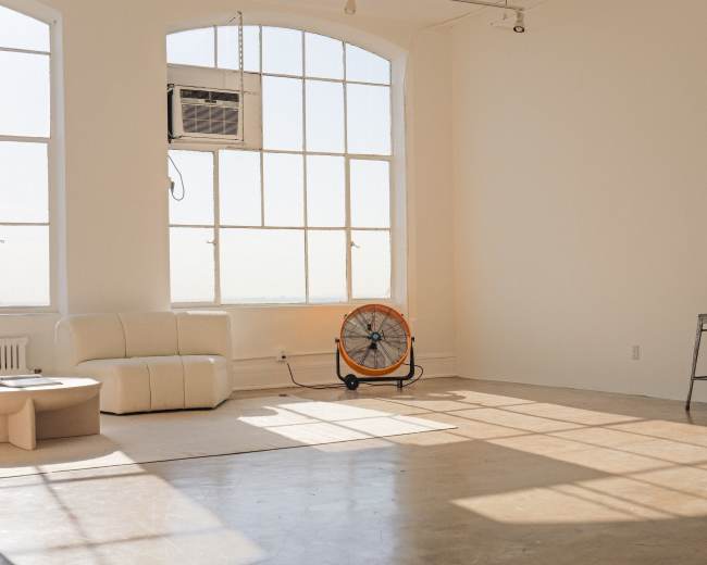 The image shows a bright, empty room with large windows, a white couch, an industrial fan, a folding stool, and a small cube-shaped table.