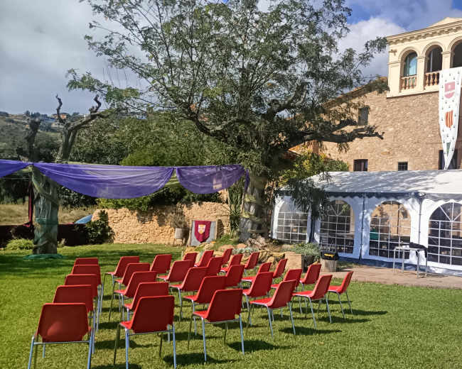 A wedding venue setup features rows of red chairs facing a large white tent beside a rustic stone building, surrounded by greenery.