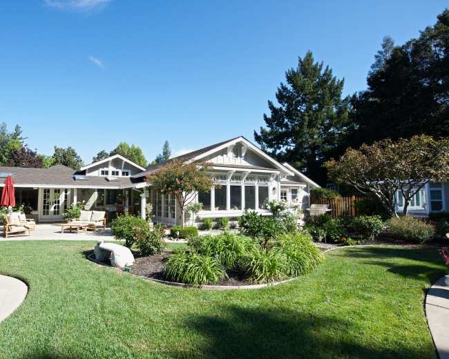 A well-maintained green lawn surrounds a large, modern single-story house with multiple windows and a covered patio area.