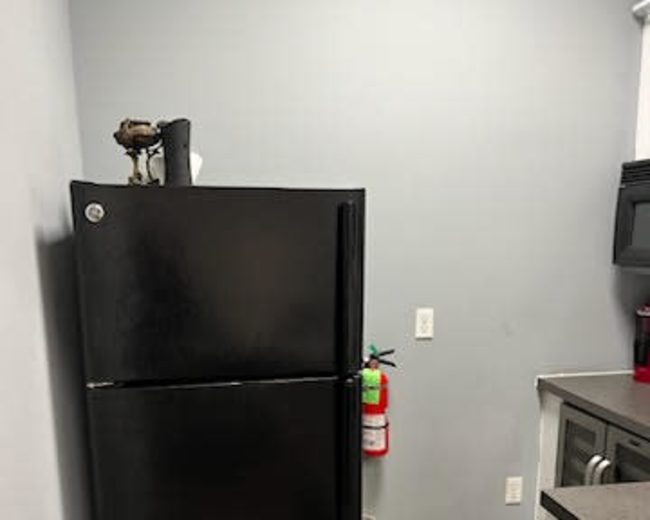 A black refrigerator stands against a gray wall in a kitchen with a fire extinguisher and trash can nearby.