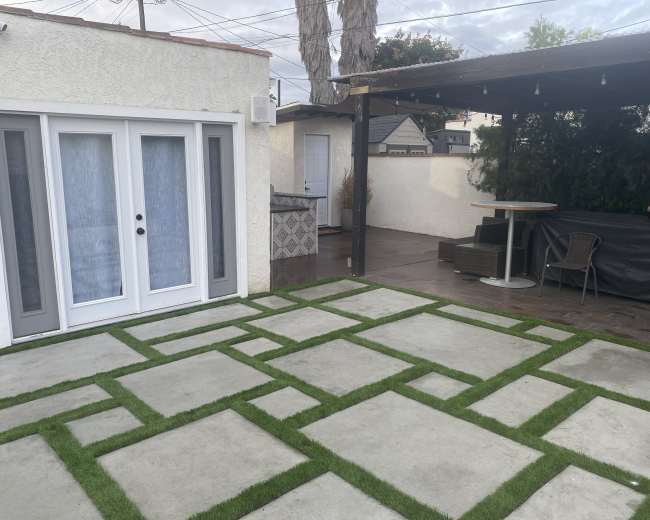 The image shows a patio area featuring a concrete and grass pattern, with an outdoor seating space and a small building in the background.