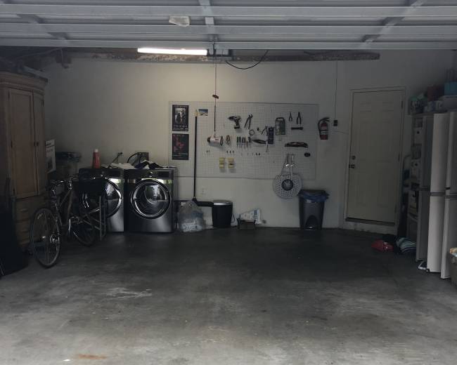 The garage contains two washing machines, a bicycle, a trash can, and various tools hanging on a wall, with a refrigerator in the back corner.