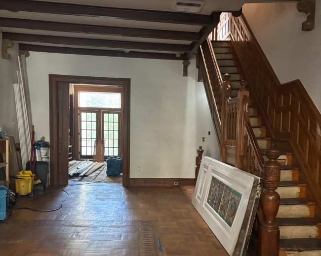 The image shows the interior of a partially furnished room with wooden flooring, a staircase leading to an upper level, and a doorway leading to another room.