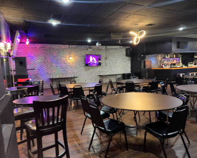 The image shows an empty restaurant or bar area with round tables, chairs, and a bar in the background, featuring brick walls and low lighting.
