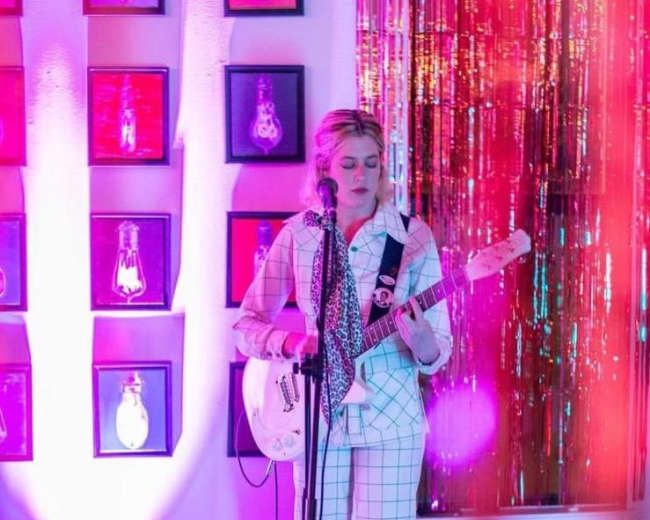 A musician stands on stage with a guitar, surrounded by colorful lights and decorations, as balloons lie on the floor.