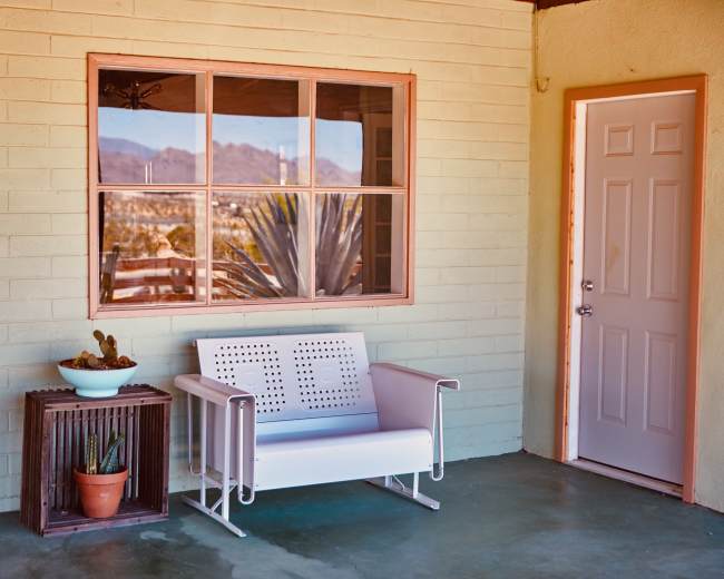 The image shows a porch area featuring a white bench, a small plant pot, and a door with a window reflecting mountains in the background.