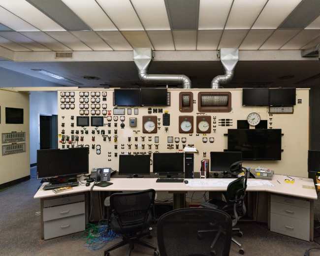 The image shows a control room with a large wall displaying various gauges, clocks, and monitors, along with a central desk equipped with multiple computer screens and chairs.