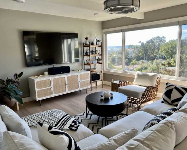 The image shows a modern living room with a large window, a television on the wall, a bookshelf, and a white sectional sofa arranged around a circular coffee table.